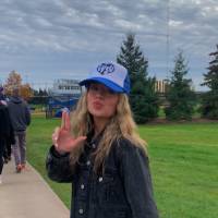 Female student holding fingers in a L-shape with a hat on, turning around